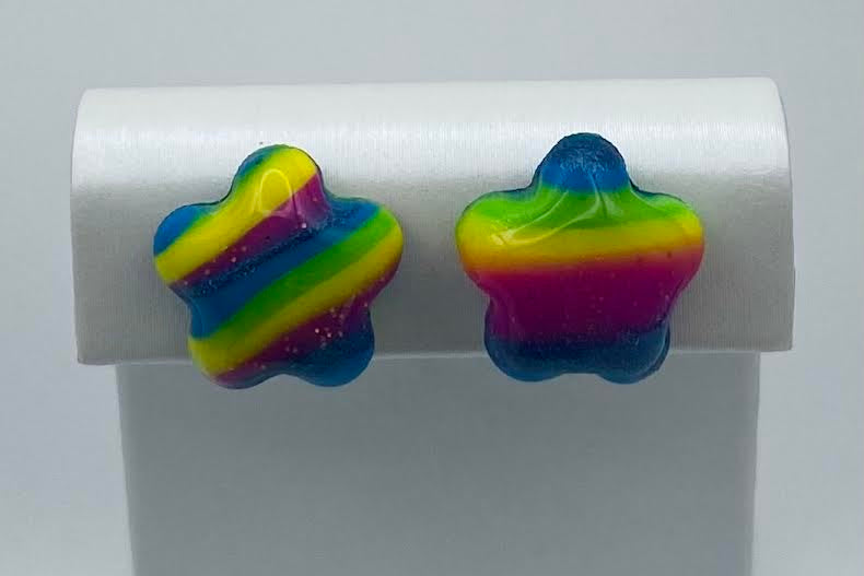 A pair of star-shaped stud earrings with a vibrant rainbow stripe pattern, displayed on a white jewelry stand against a light gray background.
