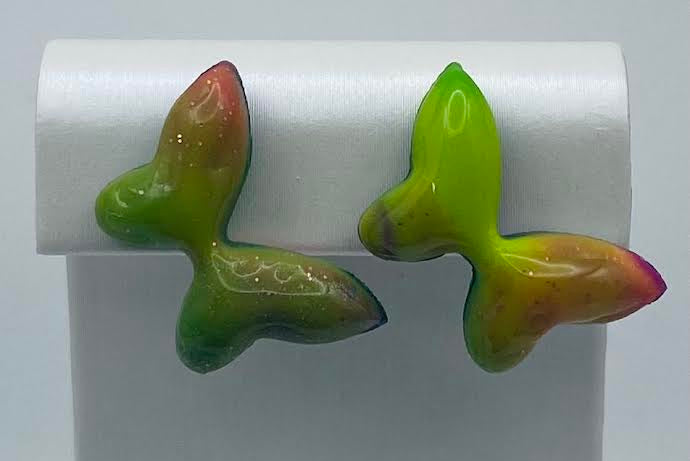 A pair of sculptural, organic-shaped earrings in a glossy green, yellow, and pink gradient finish, displayed on a white jewelry stand.