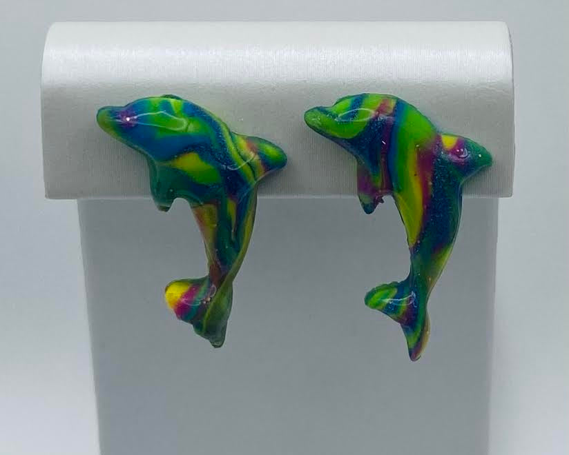 A pair of dolphin-shaped earrings with a vibrant, marbled pattern of green, blue, yellow, and purple, displayed on a white jewelry stand.