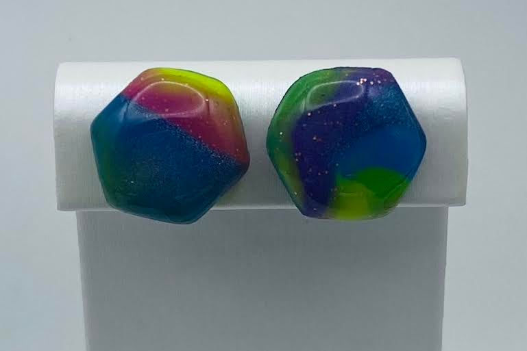 A pair of colorful hexagonal stud earrings displayed on a white stand against a light gray background. The earrings feature a marbled swirl pattern of blue, green, yellow, and pink with tiny glittery flecks.