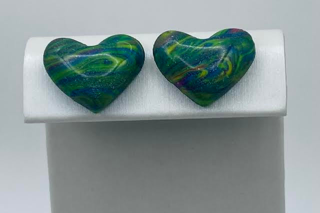 A pair of heart-shaped stud earrings with a marbled green, blue, and yellow pattern, displayed on a white jewelry stand against a light gray background.