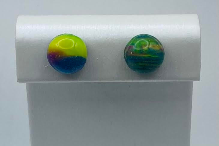 Two round, glossy stud earrings displayed on a white jewelry stand against a light gray background. One earring has a neon yellow, blue, and purple gradient design, and the other has a green and teal marbled pattern.