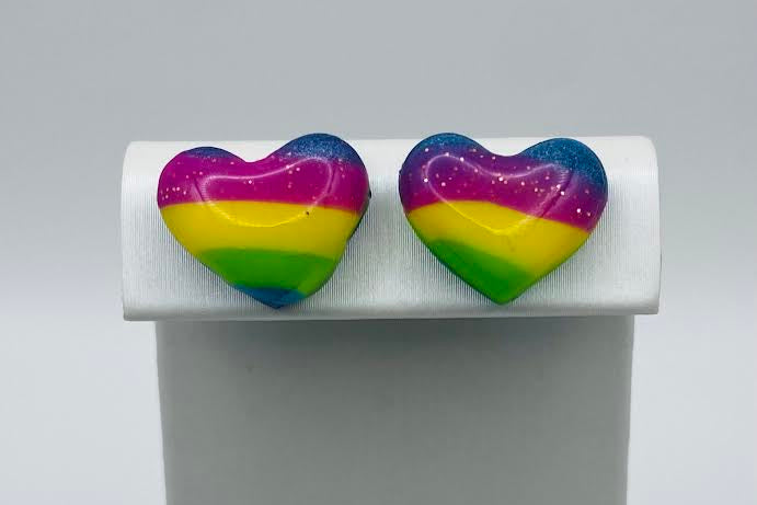A pair of heart-shaped stud earrings with a rainbow stripe pattern, displayed on a white jewelry stand against a light gray background.
