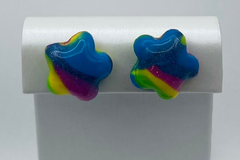 A pair of star-shaped stud earrings with a glittery rainbow gradient of blue, green, yellow, and purple, displayed on a white jewelry stand.