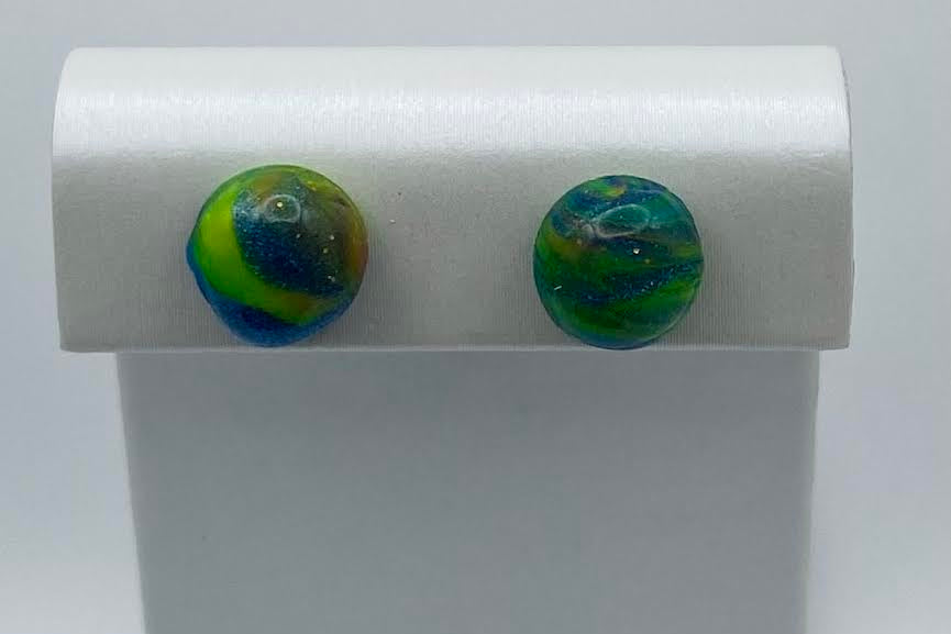 A pair of round, marbled glass stud earrings in green, blue, and yellow, displayed on a white jewelry stand against a light gray background.