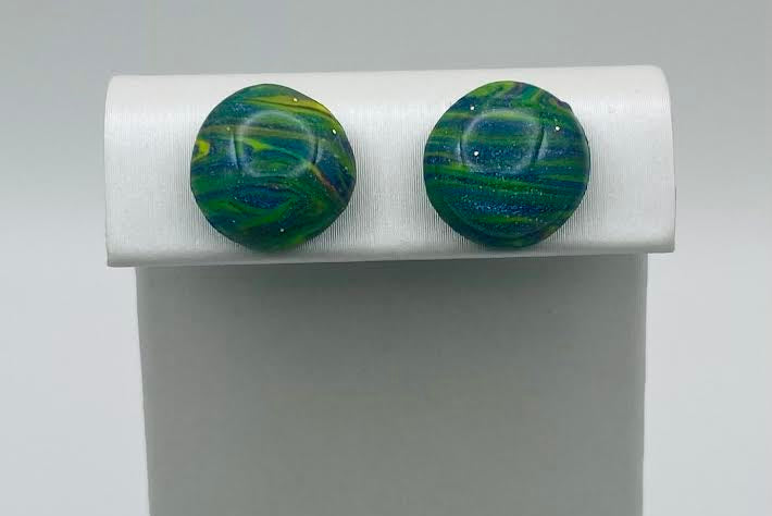 A pair of round, domed glass stud earrings with swirling green, blue, and yellow patterns, displayed on a white stand against a light gray background.
