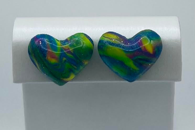 A pair of heart-shaped stud earrings with a marbled swirl pattern of green, blue, yellow, and purple, displayed on a white rectangular stand.